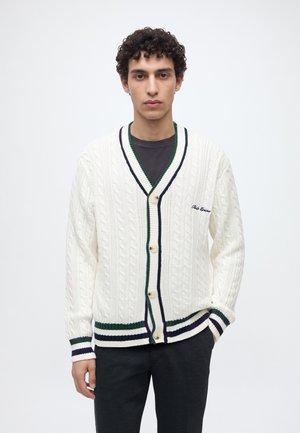 Young man with curly hair wearing a white cable-knit cardigan with green and navy stripes over a dark shirt, standing against a plain background.