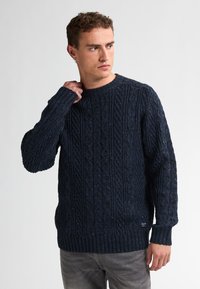 Pulover tricotat bleumarin, cu un model de cabluri, guler rotund și mansete și tiv cu dantelă. Construit dintr-un țesătură groasă și texturată.