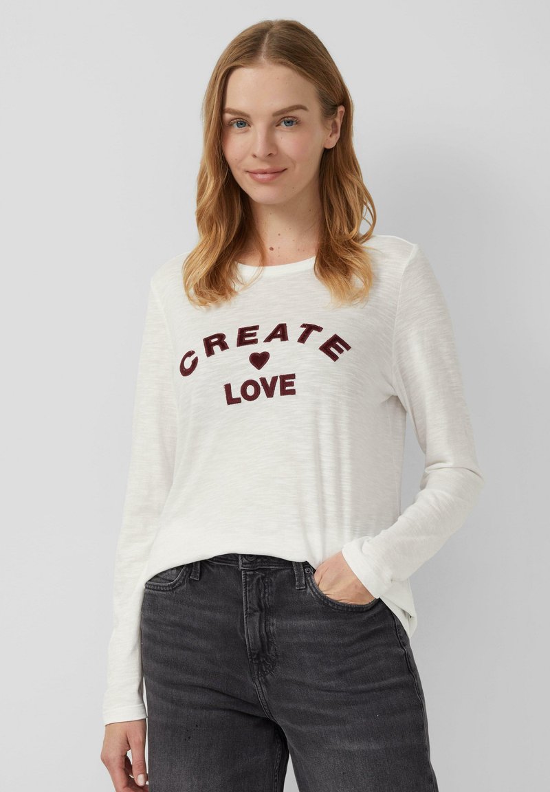 Chemise blanche à manches longues avec le texte rouge « CREATE ❤️ LOVE. » . Elle a une coupe décontractée et une texture douce, associée à un jean gris foncé.