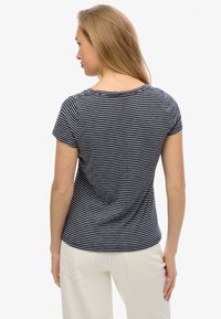 Kvinde set bagfra iført en kortærmet skjorte med navyblå og hvide striber og off-white bukser mod en ensfarvet baggrund.