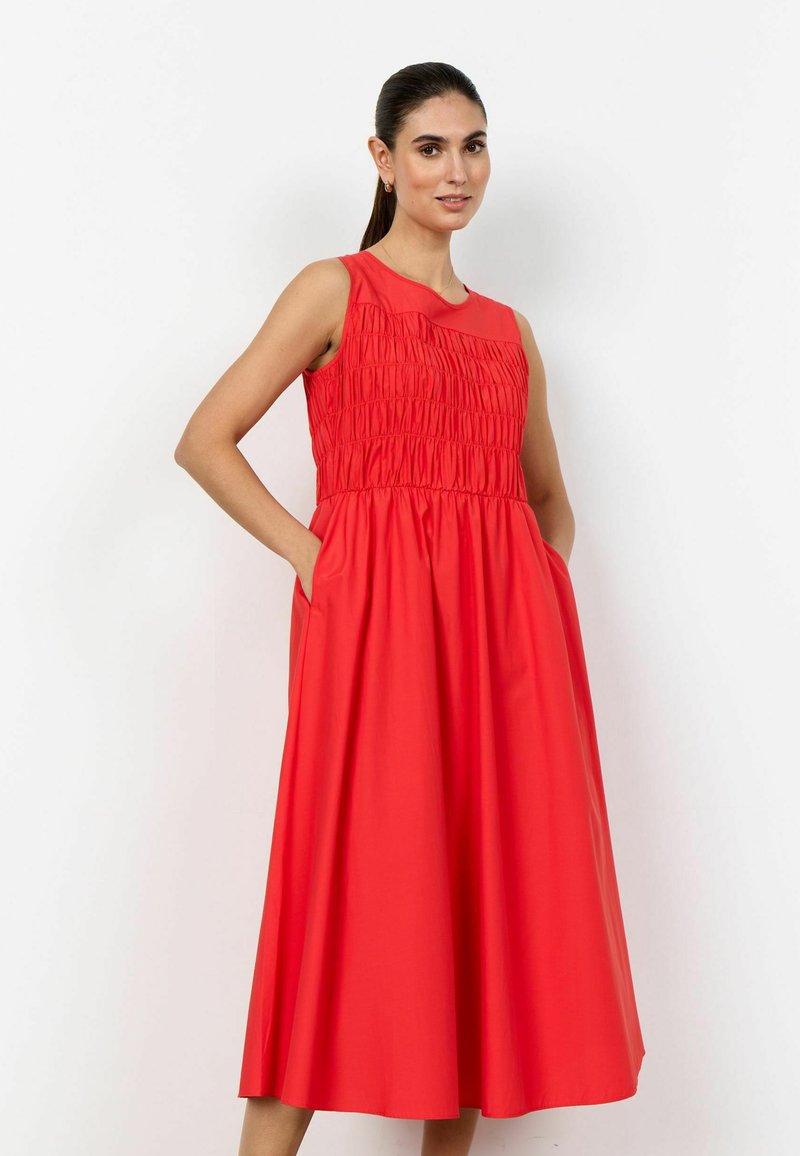 Femme aux cheveux foncés attachés en queue de cheval, portant une robe midi rouge sans manches avec un corsage froncé et les mains dans les poches de la robe, debout devant un fond blanc.