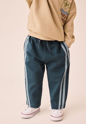 Kind trägt blaue Jogginghose mit hellblauen Seitenstreifen, beige Sweatshirt mit Bärendruck und weiße Sneakers mit rotem Detail.