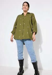 Olijfgroene overhemd met zilveren ster borduursel, knoopsluiting en opgerolde mouwen. Gecombineerd met lichtblauwe jeans en zwarte laarzen.