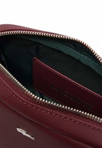 Sac texturé marron bordeaux avec fermeture éclair, dotée d'un intérieur sombre et d'une étiquette Lacoste. Quincaillerie en métal argenté et logo subtil.