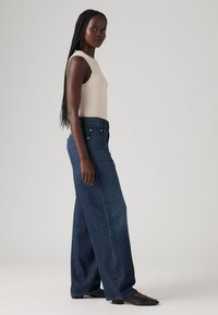 Hlače širokih nogavica od tamnog trapera s visokim strukom, u kombinaciji s bež uskim majicom bez rukava. Glatka tekstura i minimalistički dizajn.