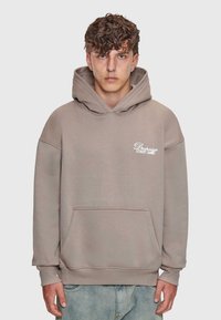 Junger Mann mit lockigem Haar trägt einen beigen Kapuzenpullover mit dem Text "Dropsize Street Game" und hellblauen Jeans, steht vor einem weißen Hintergrund.