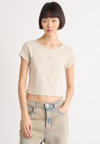 Beige avkortet t-skjorte med korte ermer, ribbet tekstur, og en liten rød og hvit logo på brystet, kombinert med lyse denimjeans.