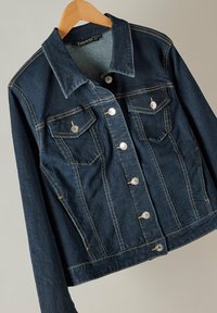 Tumma farkkutakki, jossa on kauluspanta, hopeaiset napit, kaksi rintataskua ja näkyvät ompelusarjat. Ripustettu puuhenkariin.