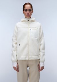 Witte fleecejas met een capuchon, gestructureerd oppervlak, ritssluiting aan de voorkant en een klein borstzakje. Gecombineerd met lichtbeige broeken.