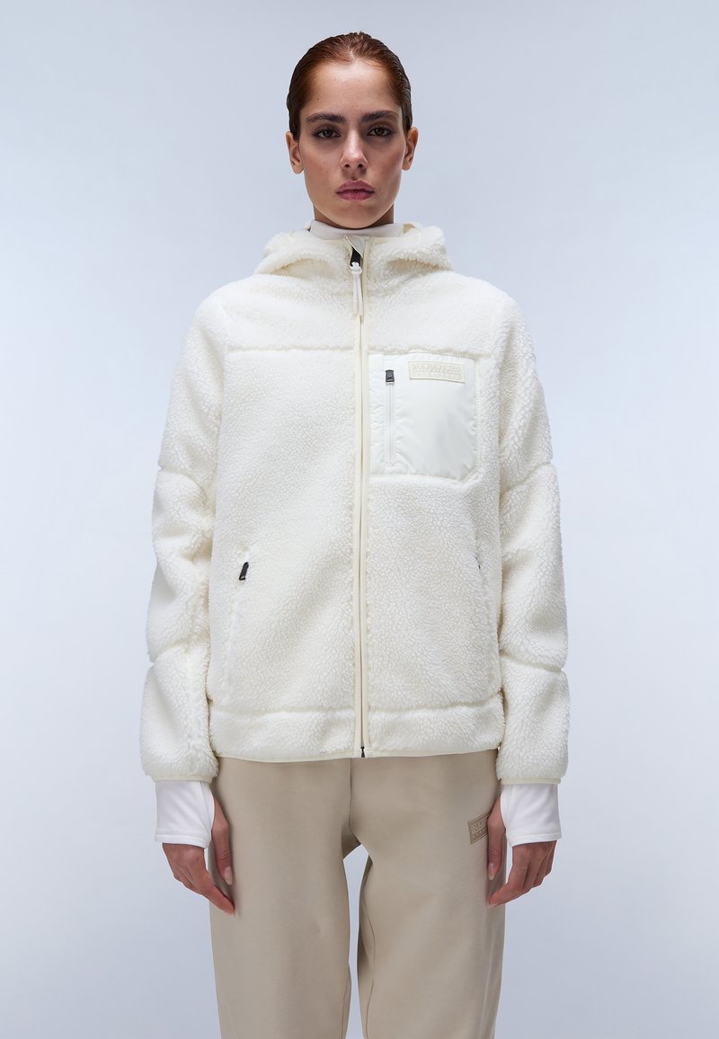 Witte fleecejas met een capuchon, gestructureerd oppervlak, ritssluiting aan de voorkant en een klein borstzakje. Gecombineerd met lichtbeige broeken.