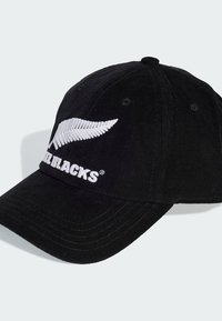 Gorra de pana negra con logotipo de helecho bordado en blanco y texto "ALL BLACKS", forma estructurada con visera curvada y ajustador de metal en la parte posterior.