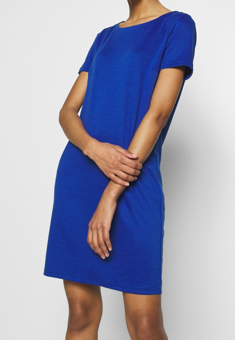 Femme portant une robe courte à manches courtes, longueur genou, bleu royal uni avec un col rond, debout les bras croisés devant elle.