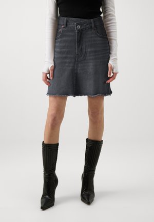 Kobieta w przyblakłej czarnej jeansowej spódnicy z postrzępionym brzegiem, białym długim rękawem, czarnej kamizelce bez rękawów i czarnych, sięgających kolan, botkach na obcasie.