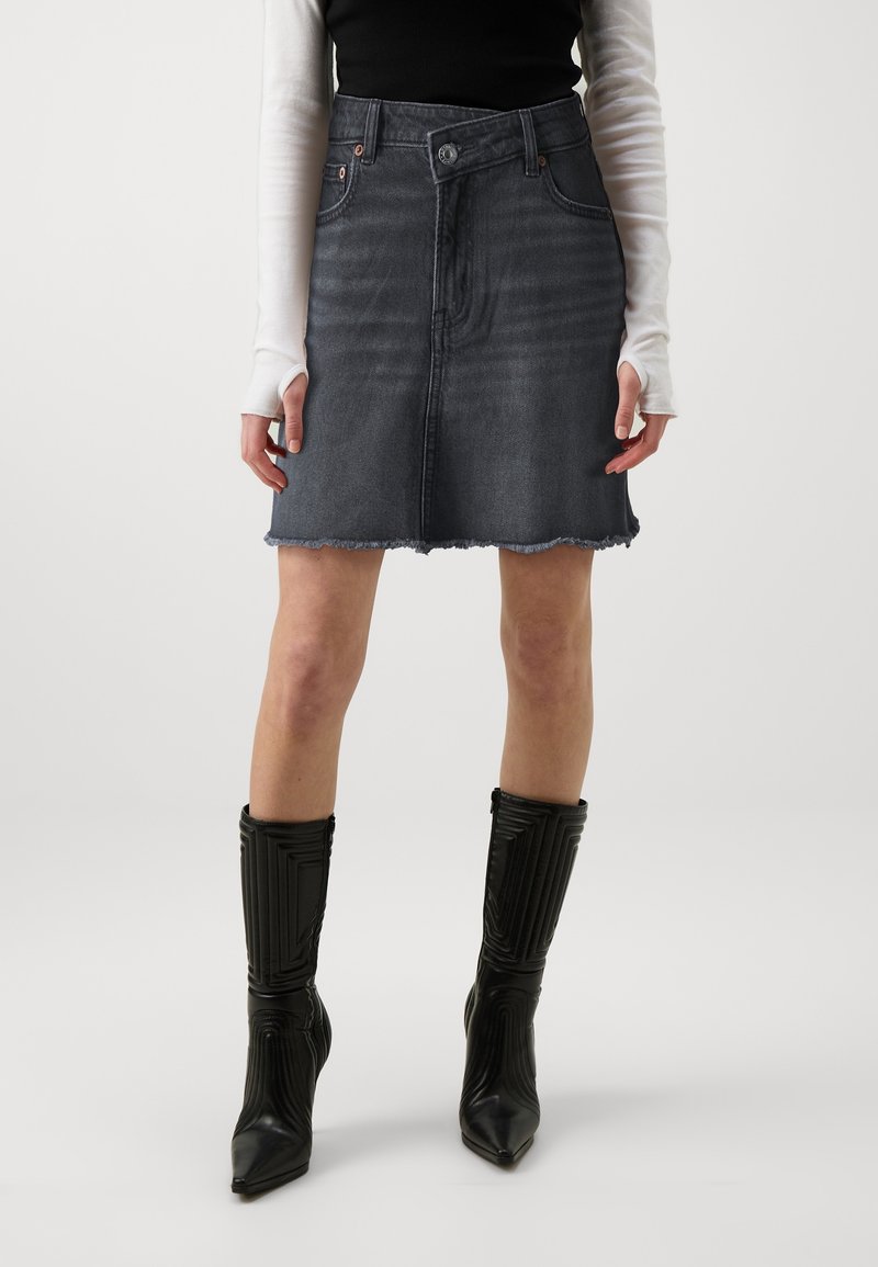 Femme portant une jupe en jean noire délavée à ourlet effiloché, un haut blanc à manches longues, un gilet noir sans manches, et des bottes noires à talons montant jusqu'aux genoux.