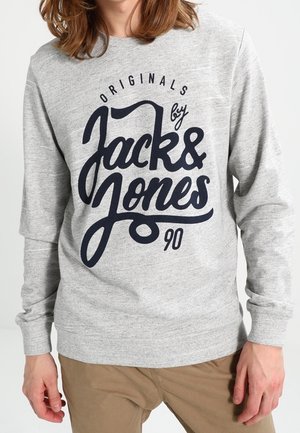 Osoba ubrana w jasnoszarą bluzę z nadrukiem "Originals by Jack & Jones 90" w dużym czarnym stylizowanym pisaniu, zestawiona z jasnobrązowymi spodniami.