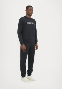 Sorte bomulds sweatshirt og matchende bukser med ribbede manchetter, med en hvid "Armani Exchange" logo på brystet. Sneaker i neutrale nuancer.