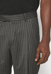 Szare spodnie w prążki z płaskim przodem, z dopasowanym pasem i widocznymi zakładkami. Materiał wydaje się mieć fakturę i może być miękki w dotyku.