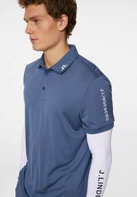 Herrblå pikétröja med korta ärmar, med en strukturerad krage, tre knappar och vit företagslogotyp på axeln och kragen.