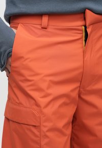 Oranžas kravas bikses no gludas auduma, ar sānu kabatu un jostas cilpu, kontrastējoša šuve redzama gar malām.