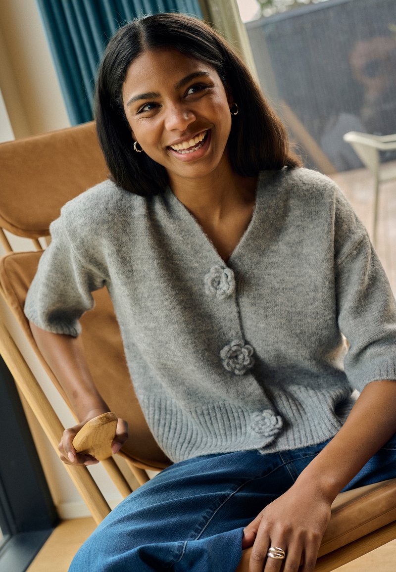 Gray knit cardigan with short sleeves, featuring three floral buttons and ribbed hem. Worn over blue jeans, held by a person on a chair.