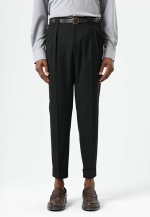 Pantalon noir taille haute en tissu lisse, avec des plis, des ourlets retroussés, porté avec une ceinture en cuir marron et des mocassins.