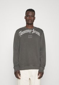 Sötét szürke pulóver, nagy fehér betűkkel a mellkason: "Tommy Jeans". Bordás mandzsettákkal és hemekkel, puha tapintással és laza szabással rendelkezik.