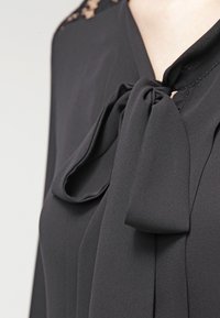 Blusa nera con un fiocco al collo, tessuto morbido, design plissettato e delicate embelliture in pizzo vicino allo scollo.