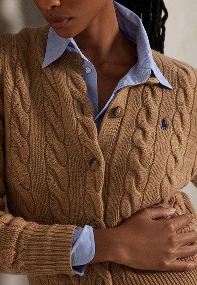 Femeie care poartă un cardigan bej cu model torsadat peste o cămașă bleu cu nasturi, cu mâna stângă sprijinită pe stomac.