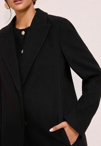 Palton negru din amestec de lână cu un design ajustat, având rever cu tăietură, două butoane și buzunare laterale cu o textură netedă.