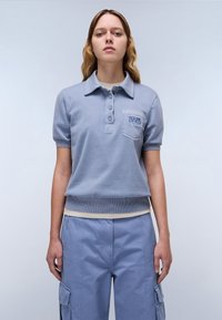 Lichtblauw poloshirt met korte mouwen, geribbelde zoom en een borstzak met logo borduring. Draagt bijpassende blauwe cargobroek.