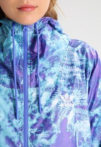 Lightweight jacket featuring a purple and blue fern-patterned design, with a zippered front and hood, accented by blue drawstrings.