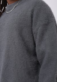 Pull gris foncé et duveteux à manches longues, porté avec un collier chaîne en argent et des cheveux tressés partiellement visibles.