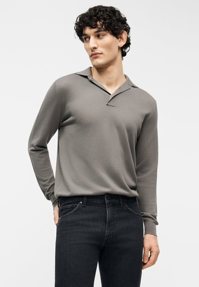 Νέος άνδρας με σγουρά μαλλιά που φοράει γκρι μακρυμάνικη μπλούζα πόλο και μαύρο τζιν, κοιτάει προς τα αριστερά με το ένα χέρι στην τσέπη.
