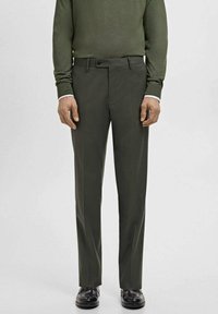 Man wearing dark green tailored trousers, matching green sweater, white shirt cuffs, and black dress shoes standing on a plain background.