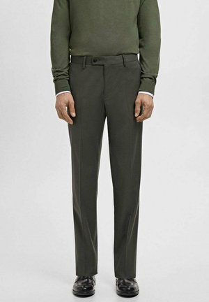 Mężczyzna ubrany w ciemnozielone, dopasowane spodnie, zielony sweter, białe mankiety koszuli i czarne eleganckie buty stojący na jednolitym tle.