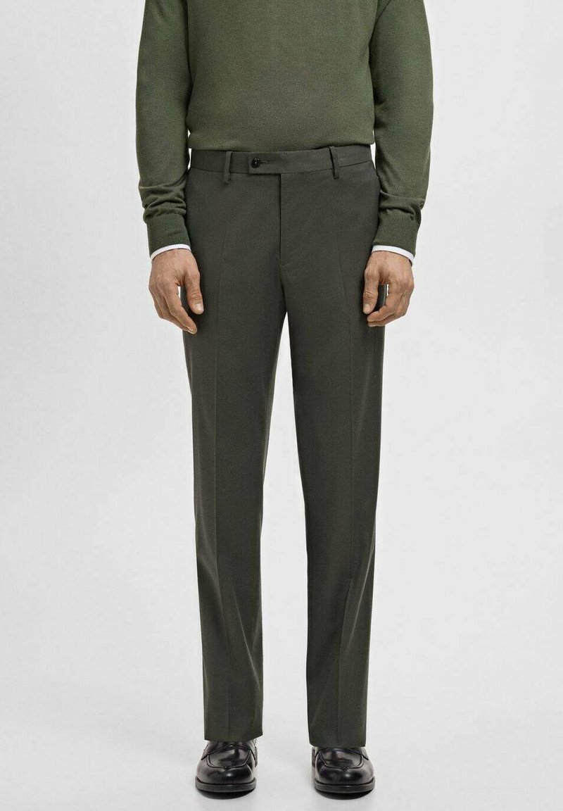 Man wearing dark green tailored trousers, matching green sweater, white shirt cuffs, and black dress shoes standing on a plain background.