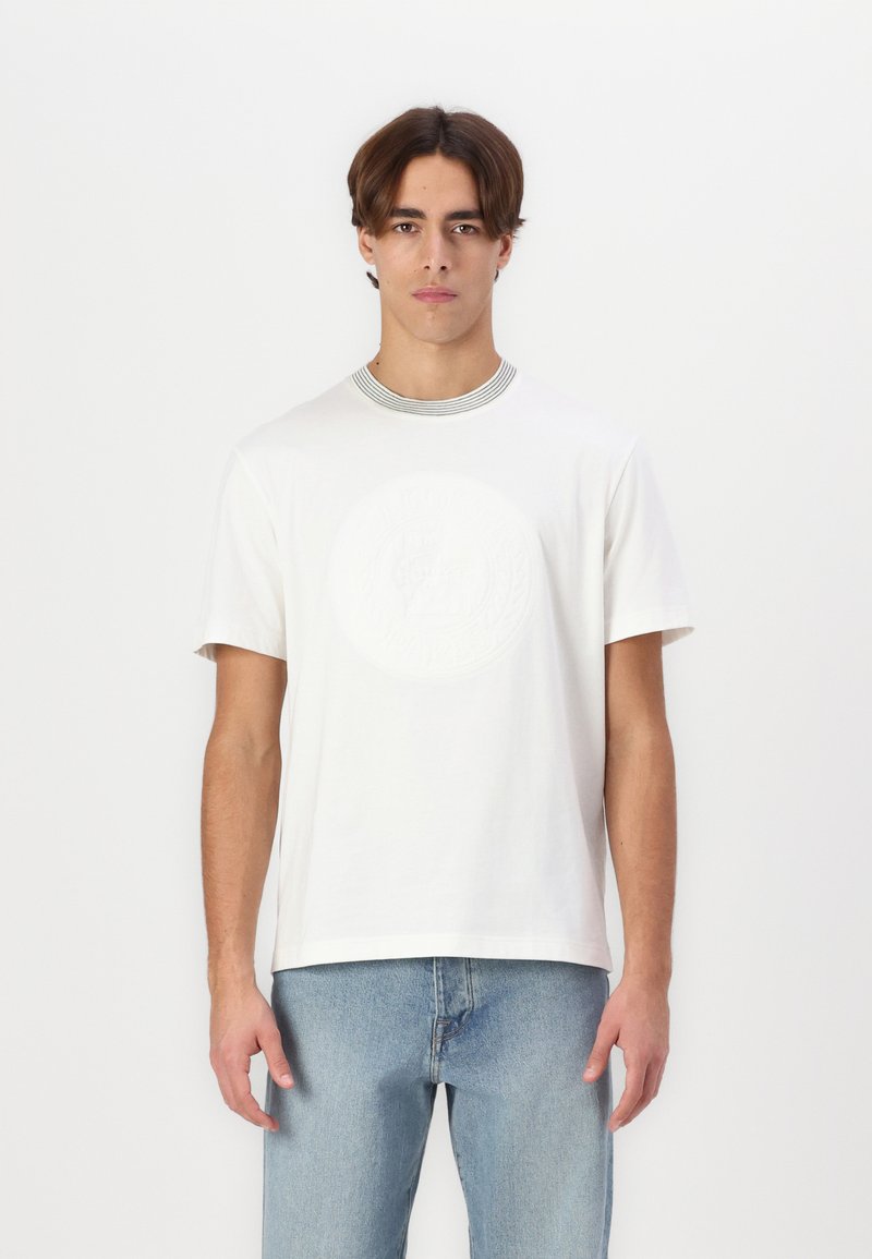 Hvid kortærmet T-shirt med et struktureret rundt logo på fronten og en stribet kraveskæring. Båret med lyseblå jeans.
