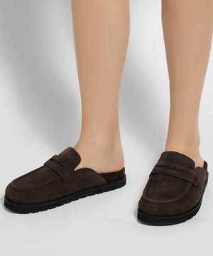 Personne portant des mocassins en daim brun foncé sans talon avec des semelles noires sur un sol clair.