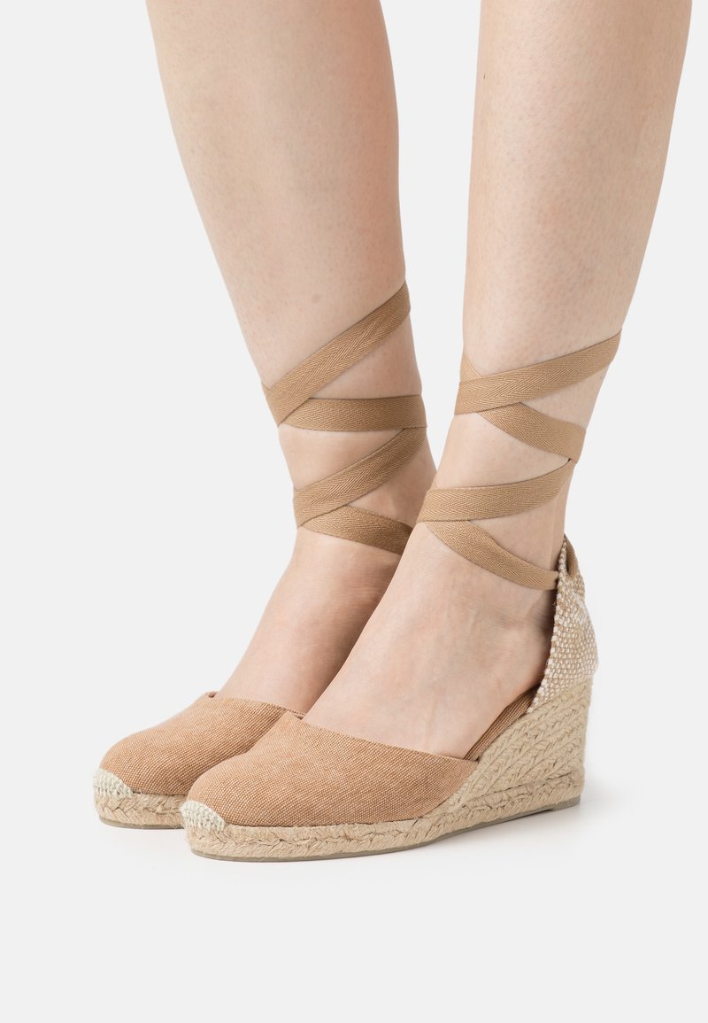 Pieds portant des espadrilles compensées beige avec des lanières nouées autour de la cheville sur un fond blanc.