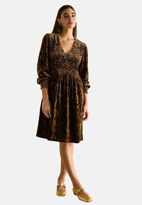 Braun gemustertes Kleid mit tiefem V-Ausschnitt, Puffärmeln und gerafftem Bund, aus weichem Stoff gefertigt. Kombiniert mit gelben Blockabsatzschuhen.