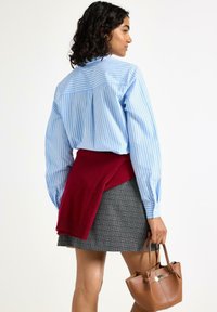 Lyseblå stribet skjorte, rød sweater bundet om livet, grå plaid nederdel og brun læderhandbag, der holdes i hånden.