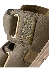 Brun läder sandal med guldfärgad hårdvara och texturerade detaljer. Designen inkluderar utskärningar och en bekväm mockainredning.