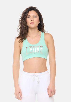 Donna con capelli castani ondulati, che indossa un reggiseno sportivo verde menta con la scritta "LEONE" e pantaloni bianchi con coulisse, in piedi davanti a uno sfondo semplice.