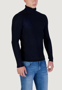 Maglione blu navy a coste con collo alto e maniche lunghe, abbinato a jeans azzurri, creando un look casual.