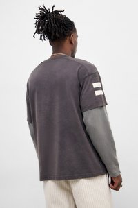 Γκρίζα μπλούζα με κοντό μανίκι, με δύο λευκές ρίγες στο αριστερό μανίκι. Φορεμένη πάνω από μια γκρι μπλούζα με μακρύ μανίκι, συνδυασμένη με ριγέ παντελόνι.