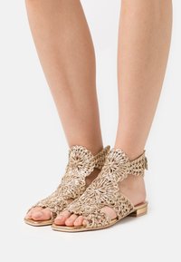 Guldcrochet-sandal med öppen tå-design, detaljerat blommönster, ankelrem och platt sula. Har en strukturerad yta och metalliska detaljer.