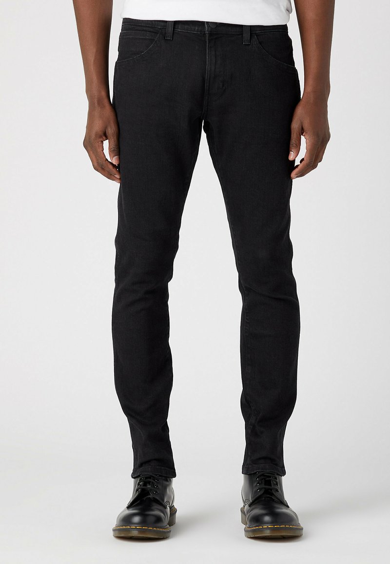 Pantalones vaqueros negros ajustados, confeccionados en denim, con cinco bolsillos, costuras visibles y un cierre clásico de botón, combinados con botas negras brillantes de cordones.