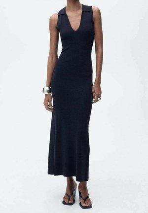 Femme portant une robe longue noire sans manches, près du corps, avec un profond décolleté en V et des sandales noires à talons hauts, debout devant un fond uni.