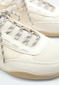 Chaussures de sport blanches avec maille texturée, lacets à motifs et semelles en caoutchouc montrées en gros plan.