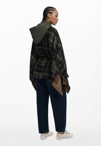 Gepunkteter Poncho mit einem dunkelgrünen Grundton, beigen und orangefarbigen Linien. Verfügt über eine kontrastierende grüne Kapuze. Getragen mit dunklen Denim-Hosen und hellen Schuhen.